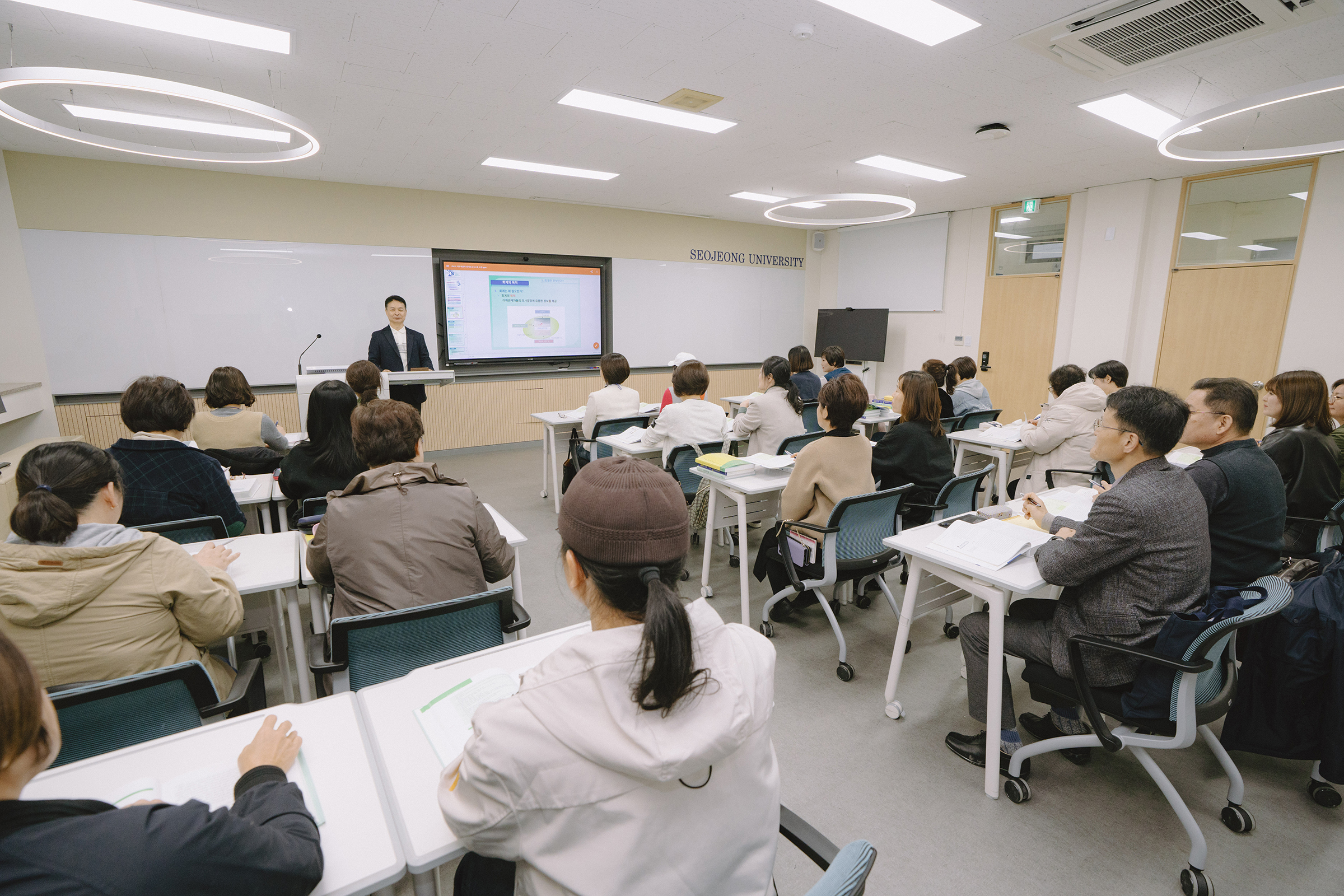 컴퓨터 실에서 수업을 듣는 학생들의 사진. 각자 모니터 앞에 앉아 집중하고 있다.